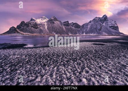 Ruhiges, gefrorenes, endloses Meer, umgeben von schwarzem, gefrorenem Sand und hohem, schneebedeckten Berg Vestrahorn bei einem malerischen Sonnenuntergang am Stockness Beach, Icel Stockfoto
