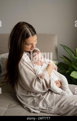 Seitenansicht einer jungen Mutter im Pyjama, die das neugeborene Baby hält, während sie morgens zu Hause auf dem Sofa sitzt Stockfoto