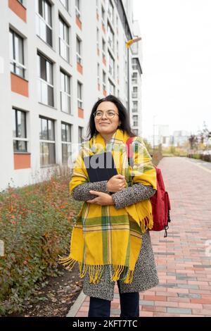 Lächelnde junge Frau in warmer Jacke und karierten Rucksack und umarmem Tablet, während sie in der Herbstsaison auf den Universitätscampus blickt Stockfoto