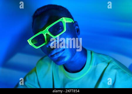 Selbstbewusster afroamerikanischer Mann mit trendigen Neon-Sonnenbrillen und grünem T-Shirt, der vor blauem Hintergrund auf die Kamera blickt Stockfoto