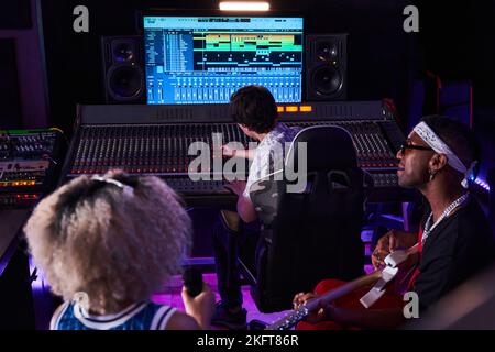 Von oben von multirassischen Menschen, die sich im Aufnahmestudio mit leuchtenden Neonlichtern versammeln, während schwarzer Mann E-Gitarre spielt und weibliche Gesänge vorsingen Stockfoto