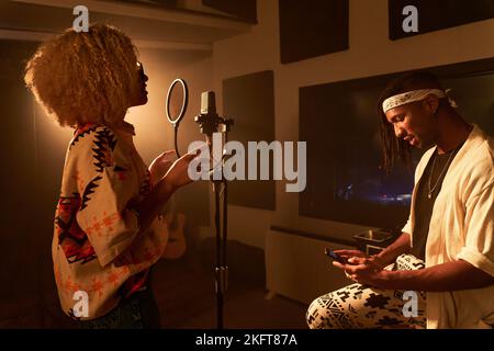 Seitenansicht eines afroamerikanischen Mannes, der sein Smartphone benutzt, während er auf einem Stuhl neben dem Mikrofon lächelt und eine Frau mit afrohaarigen Gesängen singt Stockfoto
