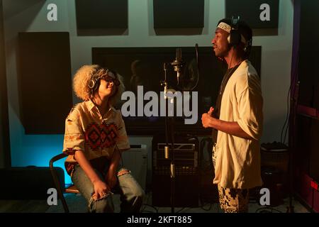 Junger afroamerikanischer Sänger steht im Studio und singt mit einem Partner, der auf einem Stuhl sitzt und Kopfhörer berührt Stockfoto