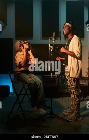 Vollkörper eines jungen afroamerikanischen Sängers, der im Studio steht und einen Song aufnimmt, mit einem Partner, der auf einem Stuhl sitzt und Kopfhörer anfasst und singt Stockfoto