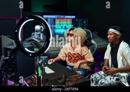 Konzentrierte, multirassische Kollegen sitzen im zeitgenössischen Aufnahmestudio mit leuchtender Neonbeleuchtung, während sie an einem neuen Song arbeiten Stockfoto