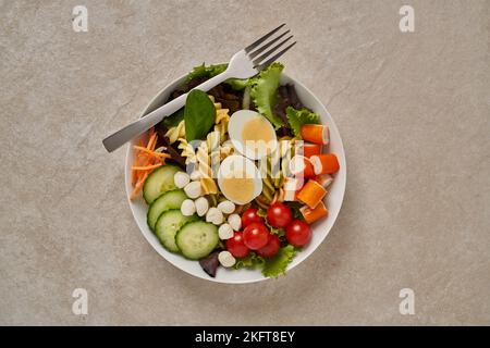 Gesunder Salat mit verschiedenen Gemüsesorten und Pasta mit gekochten Eiern und Gemüse, serviert in einer Schüssel mit Gabel auf einem Steintisch Stockfoto