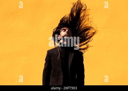 Junger hispanischer Mann in schwarzem Anzug, der die Augen schließt und lange dunkle Haare vor gelbem Hintergrund schüttelt Stockfoto