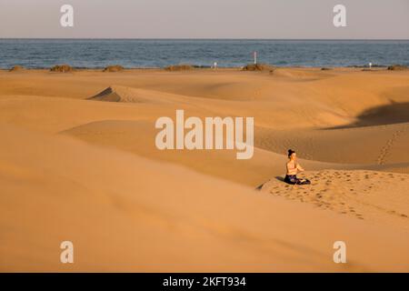 Fernansicht einer jungen Frau, die an sonnigen Tagen am Sandstrand in der Nähe des winkenden Meeres Yoga praktiziert Stockfoto