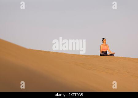 Fernansicht einer jungen Frau, die an sonnigen Tagen am Sandstrand in der Nähe des winkenden Meeres Yoga praktiziert Stockfoto