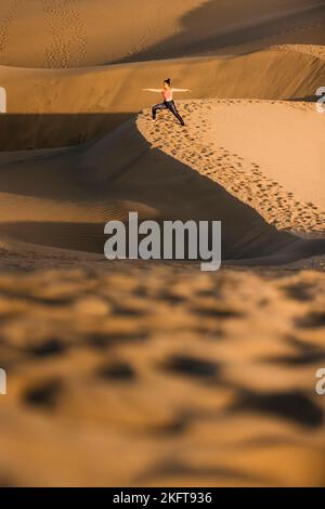 Fernansicht einer jungen Frau, die Yoga auf Sanddünen in der Wüste praktiziert. Kriegerpose. Stockfoto