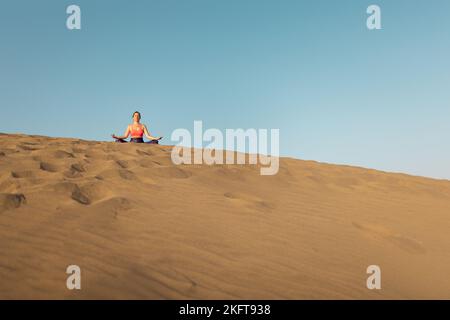 Fernansicht einer jungen Frau, die an sonnigen Tagen am Sandstrand in der Nähe des winkenden Meeres Yoga praktiziert Stockfoto