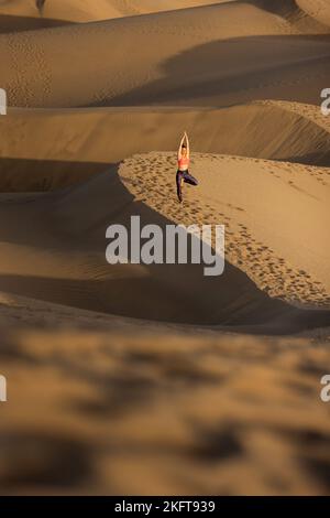 Fernansicht einer jungen Frau, die an sonnigen Tagen am Sandstrand in der Nähe des winkenden Meeres Yoga praktiziert Stockfoto