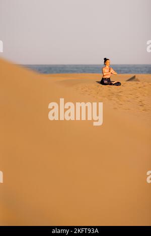 Fernansicht einer jungen Frau, die an sonnigen Tagen am Sandstrand in der Nähe des winkenden Meeres Yoga praktiziert Stockfoto