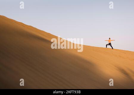 Fernansicht einer jungen Frau, die Yoga auf Sanddünen in der Wüste praktiziert. Kriegerpose. Stockfoto