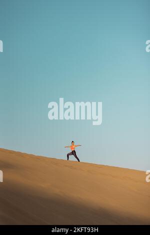 Fernansicht einer jungen Frau, die Yoga auf Sanddünen in der Wüste praktiziert. Kriegerpose. Stockfoto