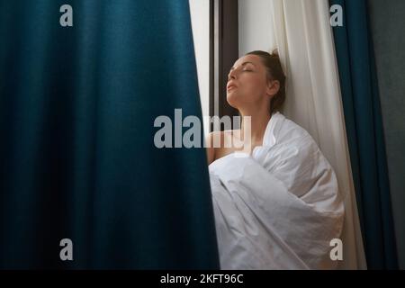 Sorglose Frau, die sich neben dem Fenster entspannt und in eine Decke gehüllt wird Stockfoto