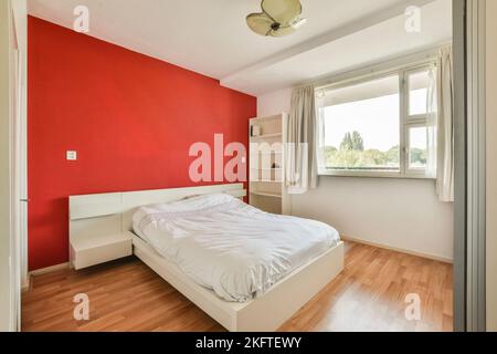 Komfortables Bett und Lampe in gemütlichem Schlafzimmer mit roten Wänden und grauen Vorhängen am Fenster in moderner Wohnung platziert Stockfoto