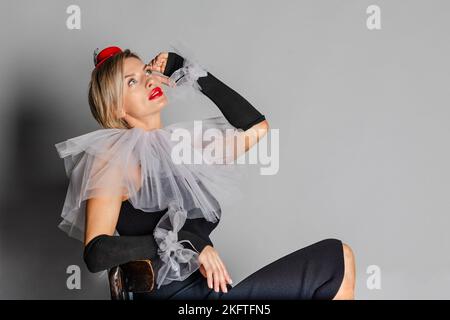 Eliteclown. Portrait einer Frau, die als Clown-Kleid mit Harlekin-Kragen und rotem Hut gekleidet ist. Kontrastbild auf grauem Hintergrund. Hochwertige Fotos Stockfoto