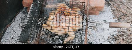 Hühnerfleischfutter werden auf dem Grill auf schwelenden Kohlen oder vom Lagerfeuer am Boden am Sommer- oder Herbsttag gebraten. Grill in Campingbedingungen. Sur Stockfoto