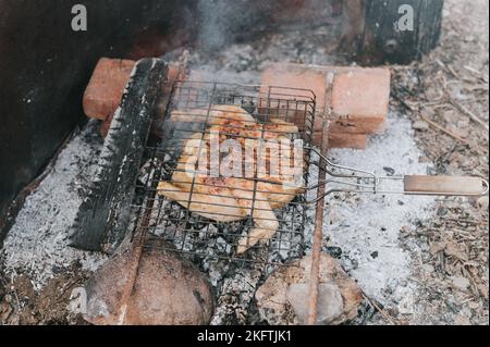 Hühnerfleischfutter werden auf dem Grill auf schwelenden Kohlen oder vom Lagerfeuer auf dem Boden am Sommer- oder Herbsttag gebraten. Grill im Campingzustand Stockfoto