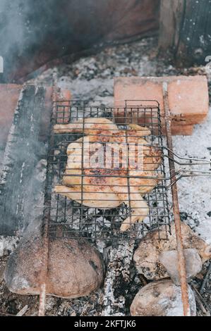 Hühnerfleischfutter werden auf dem Grill auf schwelenden Kohlen oder vom Lagerfeuer auf dem Boden am Sommer- oder Herbsttag gebraten. Grill im Campingzustand Stockfoto