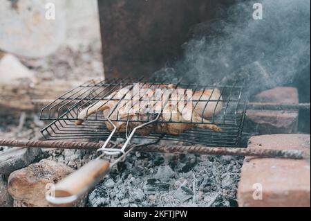 Hühnerfleischfutter werden auf dem Grill auf schwelenden Kohlen oder vom Lagerfeuer auf dem Boden am Sommer- oder Herbsttag gebraten. Grill im Campingzustand Stockfoto
