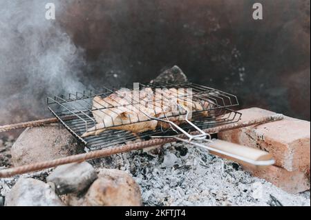 Hühnerfleischfutter werden auf dem Grill auf schwelenden Kohlen oder vom Lagerfeuer auf dem Boden am Sommer- oder Herbsttag gebraten. Grill im Campingzustand Stockfoto