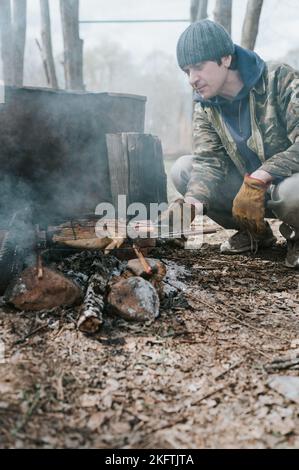 Junger Mann Survivalist kocht Braten Hühnerfleisch Lebensmittel werden auf Grill auf schwelenden Kohlen oder Ember von einem Lagerfeuer auf dem Boden gebraten. Grill im Campingplatz Stockfoto