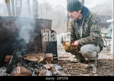 Junger Mann Survivalist kocht Braten Hühnerfleisch Lebensmittel werden auf Grill auf schwelenden Kohlen oder Ember von einem Lagerfeuer auf dem Boden gebraten. Grill im Campingplatz Stockfoto