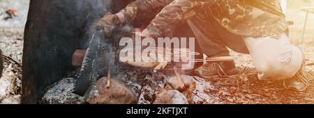 Mann Survivalist kocht Braten Huhn Fleisch Lebensmittel werden auf dem Grill auf schwelende Kohlen oder Emder vom Lagerfeuer auf dem Boden gebraten. Grill in Campingbedingungen Stockfoto