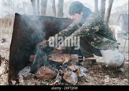 Junger Mann Survivalist kocht Braten Hühnerfleisch Lebensmittel werden auf Grill auf schwelenden Kohlen oder Ember von einem Lagerfeuer auf dem Boden gebraten. Grill im Campingplatz Stockfoto