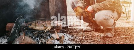 Mann Survivalist kocht Braten Huhn Fleisch Lebensmittel werden auf dem Grill auf schwelende Kohlen oder Emder vom Lagerfeuer auf dem Boden gebraten. Grill in Campingbedingungen Stockfoto