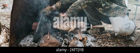 Junger Mann Survivalist kocht Braten Huhn Fleisch Lebensmittel werden auf dem Grill auf schwelende Kohlen oder Emder vom Lagerfeuer auf dem Boden gebraten. Grill in Camping Cond Stockfoto