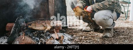 Junger Mann Survivalist kocht Braten Huhn Fleisch Lebensmittel werden auf dem Grill auf schwelende Kohlen oder Emder vom Lagerfeuer auf dem Boden gebraten. Grill in Camping Cond Stockfoto