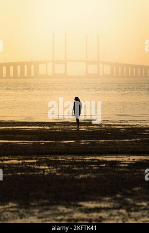 Die Silhouette einer Frau, die während eines strahlend gelben Sonnenuntergangs am Strand steht, mit einer Brücke im Hintergrund Stockfoto