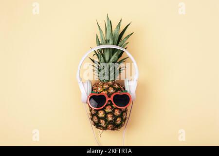 Ananas in Sonnenbrille mit Kopfhörern auf hellgelbem Hintergrund. Sommerkonzept. Draufsicht, flach liegend. Stockfoto