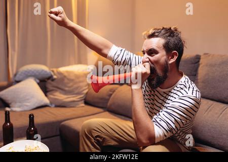 Junger begeisterter Fußballfan, der auf dem Sofa sitzt und seine Nationalmannschaft im Fernsehen jubelt Stockfoto