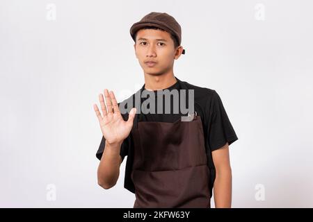 Hübscher Barista asiatischer Mann mit brauner Schürze und schwarzem T-Shirt isoliert auf weißem Hintergrund ernster Ausdruck dabei Stop-Zeichen mit Handfläche des Ha Stockfoto