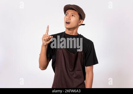 Ein hübscher, asiatischer Barista mit brauner Schürze und schwarzem T-Shirt auf weißem Hintergrund, der mit dem Finger auf dem Kopf denkt. Habe eine Idee Lösung Kontra Stockfoto