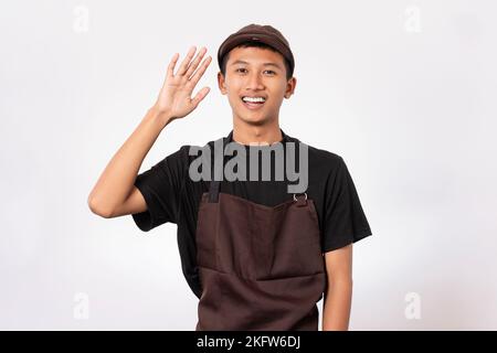 Ein hübscher, asiatischer Barista, der eine braune Schürze und ein schwarzes T-Shirt auf weißem Hintergrund trägt, lächelt freundlich und bietet Handshake und Welco Stockfoto