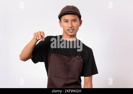 Ein hübscher, asiatischer Barista mit brauner Schürze und schwarzem T-Shirt, isoliert auf weißem Hintergrund, ärgerte sich ärgerlich, aggressiv zu sein Stockfoto