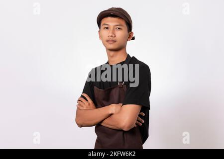 Ein hübscher, asiatischer Barista mit brauner Schürze und schwarzem T-Shirt auf weißem Hintergrund, der mit gekreuzten Armen und einem lächelnden, selbstbewussten Look steht Stockfoto