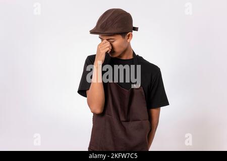 Ein hübscher, asiatischer Barista mit brauner Schürze und schwarzem T-Shirt auf weißem Hintergrund. Barista berührt den Kopf und leidet unter Kopfschmerzen, Verzweiflung Stockfoto