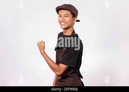 Ein hübscher, asiatischer Barista, der eine braune Schürze und ein schwarzes T-Shirt auf weißem Hintergrund trägt, das Muskelbizeps, eine Passform und ein gesundes Konzept zeigt Stockfoto