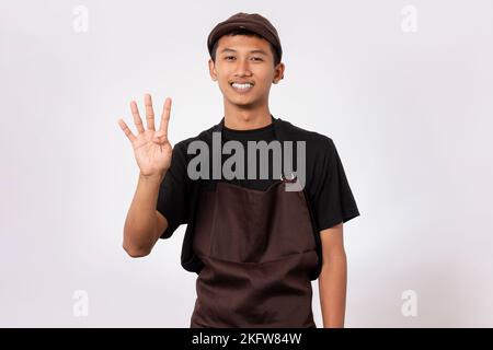 Ein hübscher, asiatischer Barista, der eine braune Schürze und ein schwarzes T-Shirt auf weißem Hintergrund trägt und mit den Fingern eine Zahlengeste macht Stockfoto