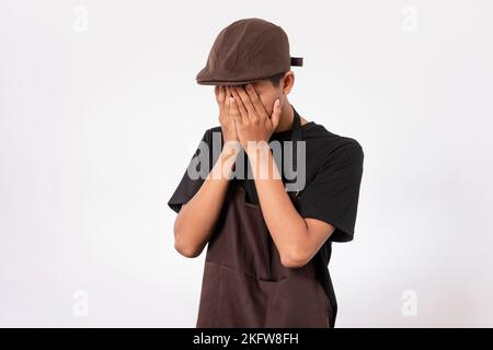 Schöner, asiatischer Barista mit brauner Schürze und schwarzem T-Shirt auf weißem Hintergrund, trauriger und müder Gesichtsausdruck. Gesicht mit Händen abdecken Stockfoto