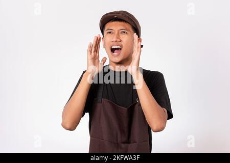 Ein hübscher, asiatischer Barista mit brauner Schürze und schwarzem T-Shirt auf weißem Hintergrund, der laut schrie und schrie. Lautes Verkaufsgeschrei ausrufen Stockfoto