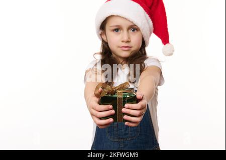 Schönes kleines Mädchen, ein kleines Kind in Weihnachtsmütze und E-Denim Overalls, überreicht ein glückliches Weihnachtsgeschenk und schaut in die Kamera Stockfoto