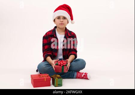 In voller Länge Porträt: Schöner präteen Junge in Santa Hut, halten Weihnachtsgeschenk, sitzt auf weißem Hintergrund. Speicherplatz kopieren Stockfoto