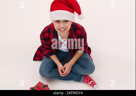Blick von oben auf einen entzückenden Teenager-Jungen im Weihnachtsmütze, der auf weißem Hintergrund mit Weihnachtsgeschenken sitzt. Speicherplatz kopieren Stockfoto